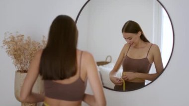 Slimming and dieting result. Young happy sporty woman checking her body condition, measuring her waist with tape and enjoying result, looking at mirror, tracking shot, slow motion, empty space