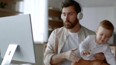 Remote work and parenthood. Young busy father businessman video chatting with partners via pc and carrying his active newborn baby on hands, working from home, tracking shot, free space