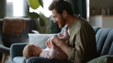 Carefree fatherhood. Young loving dad playing with his adorable laughing baby girl lying on his knees, babysitting at home, tracking shot, slow motion, free space