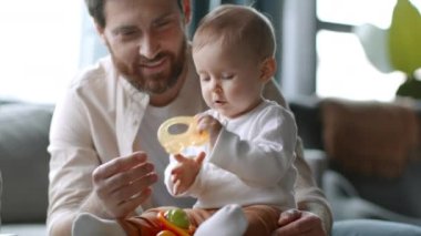 Happy childhood. Carefree little baby girl playing with toy and teether at home, loving dad sitting near and laughing, close up portrait