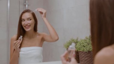 Hairstyle treatment. Young pretty woman applying nourishing serum on head skin, curing her hair at home, looking at mirror at bathroom, over shoulder view, tracking shot, slow motion, free space