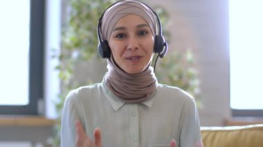 Online mentoring concept. Camera pov portrait of young positive middle eastern woman life coach wearing headset talking to camera, recording video blog at home