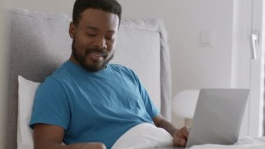 Comfortable work at home. Young carefree african american guy working on laptop in bed, answering phone call and talking with friend, resting in morning, tracking shot, slow motion, free space