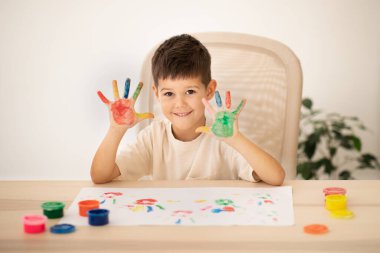 Happy small european kid with colorful hands palms enjoys drawing at table with paint in white children room interior. Art and fantasy, entertainment and fun, education, dreams and childhood at home