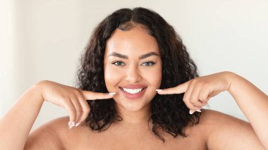 Oral care concept. Beautiful plus size woman pointing at her perfect smile with two fingers, showing white teeth, standing in bathroom, panorama
