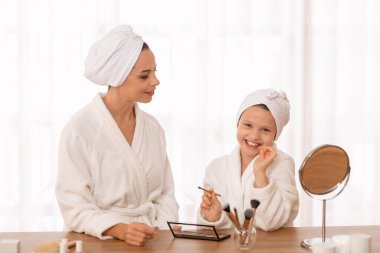 Beautiful mother and her little daughter in bathrobes doing makeup at home, happy young mom and preteen female child having beauty day, testing new cosmetics, enjoying spending time together