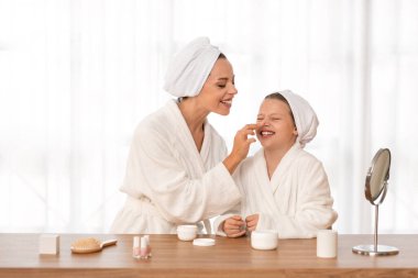 Happy mother and little daughter in bathrobes and towels applying face cream while having beauty day together at home, young mom playfully touching nose of her preteen female child and laughing