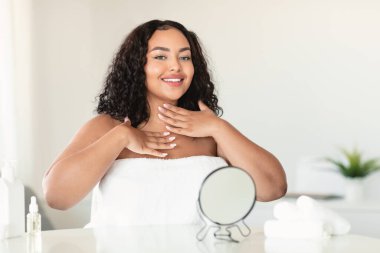 Perfect healthy skin. Happy body positive woman in towel applying moisturizing cream on her neck, looking and smiling at camera. Plus size lady using natural cosmetic product for her body
