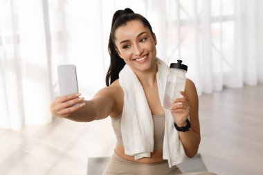 Pretty cheerful smiling brunette young woman in sportswear fitness blogger taking selfie on cell phone, athletic lady yoga instructor showing bottle of water, recommend water drinking during workout