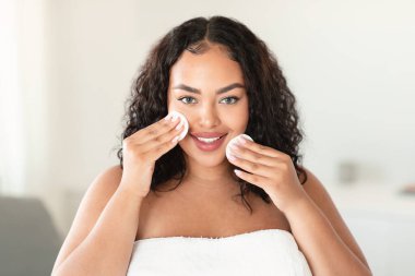 Portrait of excited plus size woman using cotton pads, erasing make up from her face, applying facial toner or cleansing milk, looking and smiling at camera, copy space