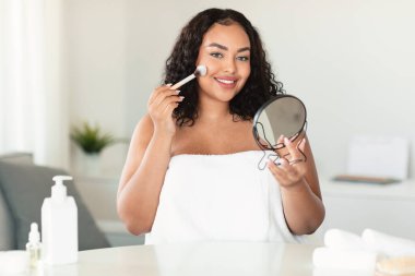 Makeup, visage concept. Bodypositive lady holding mirror, applying blush and smiling at camera, home interior, copy space. Attractive woman applying nude makeup, using foundation and brush