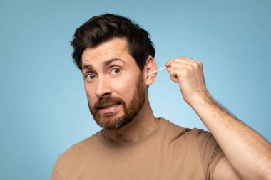 Funny middle aged bearded man cleaning ears with cotton swab, doing daily hygiene routine, blue studio background. Clean ear and safe earwax removal