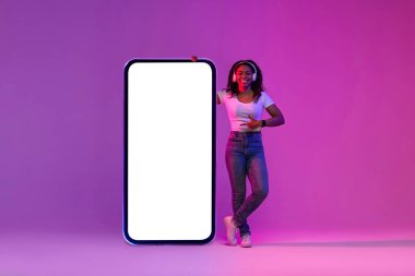 Cheerful African American Woman In Headphones Pointing At Big Blank Smartphone With White Screen, Smiling Black Female Showing Copy Space For App Or Website Design, Standing In Purple Neon Light