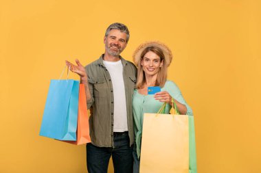 Easy payment. Excited middle aged couple showing debit credit card and holding shopper bags after purchases, standing over yellow studio background