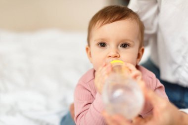 Closeup shot of cute little infant girl drinking fresh water from bottle, woman holding daughter and helping kid, sitting on bed, free copy space