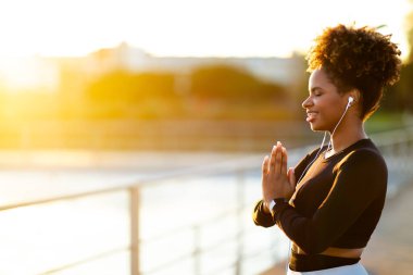Yoga Outdoors. Portrait Of Smiling Young Black Woman Meditating Outside In The Morning, Sporty Female Making Namaste Gesture With Closed Eyes And Listening Music In Earphones, Copy Space