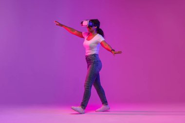Excited Black Lady Wearing Modern VR Headset Experiencing Virtual Reality In Neon Light, Happy Young African American Female Walking On Purple Background And Touching Air While Playing Video Game