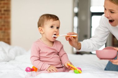 Breakfast for kid. Mother feeding her adorable baby daughter with porridge, sitting on bed in bedroom interior, free space. Child opening mouth and eating healthy food