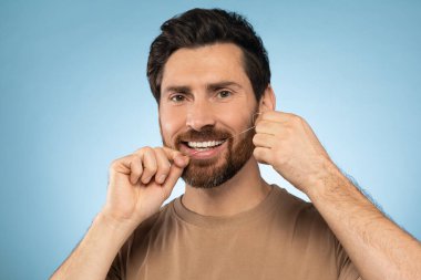 Happy middle aged man using dental thread floss and smiling, cleaning teeth after food posing over blue background, studio shot. Dental care concept