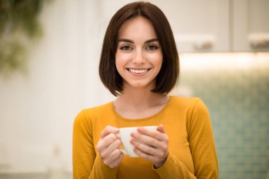 Portrait of happy cheerful pretty young woman in casual enjoying fresh aromatic coffee, holding mug and smiling at camera, lady drinking herbal tea in the morning, modern kitchen interior, copy space