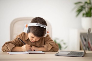 Busy small european boy in headphones sitting at table, studying, drawing in room interior. Lesson and school, art, learning at home and children garden with device, childhood and education remotely
