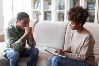 Beautiful millennial black woman with bushy hair psychologist have conversation with frustrated unhappy pensive young african american man, therapist looking at patient, taking notes, copy space