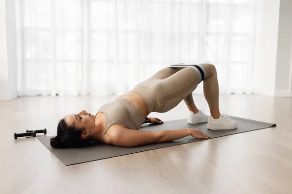 Healthy lifestyle concept. Well-fit sporty young woman exercising at home, athletic lady in beige sportswear lying on yoga mat, doing morning workout, using fitness tools, copy space, full length