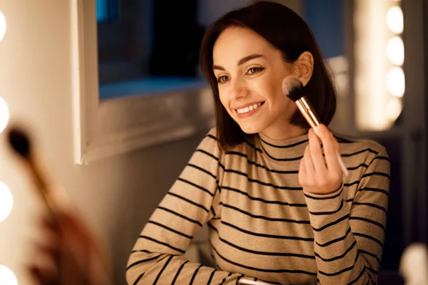 Makeup, visage concept. Cheerful pretty young brunette woman looking at mirror, applying blush, home interior. Attractive lady applying natural light makeup, using foundation and brush, going out