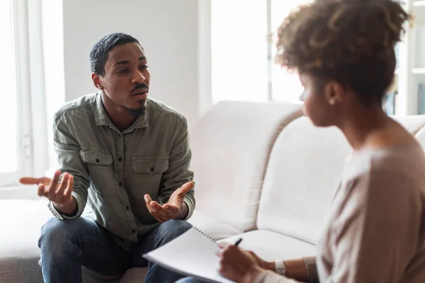 Unhappy anxious depressed young black man attending therapy session with psychologist, male patient sitting on couch at counselor office, sharing his situation with woman therapist, copy space