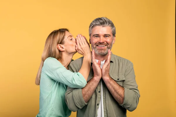 Gossip, share advice. Woman whispering to her husband on ear, happy surprised man listening wife, posing over yellow studio background