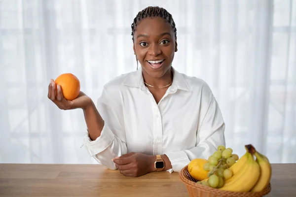 Happy african american lady hold orange in hand and smiling at camera, sitting at table with various fruits in a plate. Young healthy black woman enjoying healthy lifestyle and diet full of vitamins