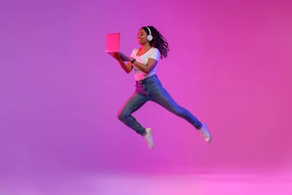 Happy Black Woman In Wireless Headphones Jumping With Laptop In Neon Light, Cheerful Young African American Lady Working Online Or Playing Video Games On Computer, Enjoying Modern Technologies
