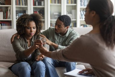Young black couple visit family psychologist together, attentive african american man embracing his anxious wife or girlfriend, spouses sitting on couch at counselor office. Crisis in relationships