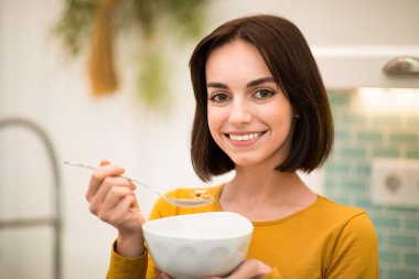 Closeup shot of cheerful smiling pretty brunette young woman enjoying healthy breakfast at home, eating cereals, whole grain granola, oatmeal, copy space. Nutrition, diet concept