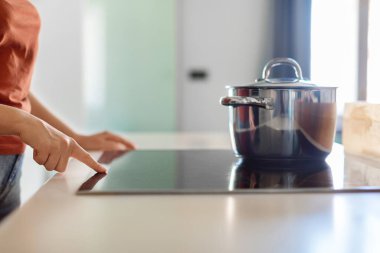 Female hand turning on induction stove with steel cooking pan in kitchen, closeup shot of woman finger touching sensor button on electrical hob while preparing food at home, side view, cropped