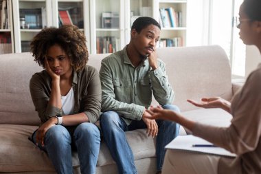 Frustrated married young black couple attending family therapist, man and woman sitting on couch back to back, avoid looking at each other while listening to female psychologist