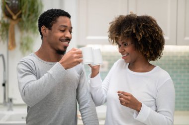 Loving millennial black spouses man and woman in homewear cheering with white coffee mugs, celebrating new day, happy couple have breakfast together, cozy kitchen interior