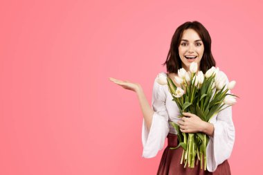 Glad emotional young caucasian lady in casual enjoy gift of bouquet of flowers show hand to free space isolated on pink background, studio. Surprise, celebration holiday, sale, ad, offer and lifestyle