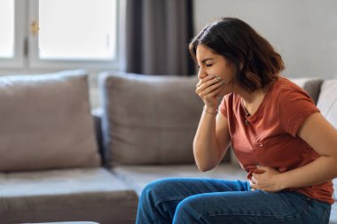 Portrait Of Young Arab Woman Suffering From Nausea At Home, Millennial Middle Eastern Female Having Morning Sickness, Ill Lady Sitting On Couch In Living Room, Covering Mouth And Touching Belly