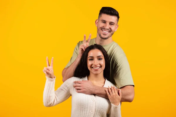 Smiling young arabic couple in casual hugging, showing peace sign with fingers and make horns, have fun, isolated on yellow background, studio. Love, romance, relationship and photo, ad and offer