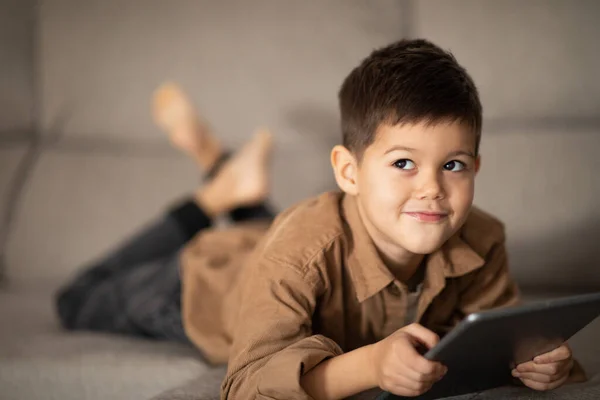 Pensive Cheerful Small European Boy Lies Sofa Thinks Tablet Look —  Fotos de Stock