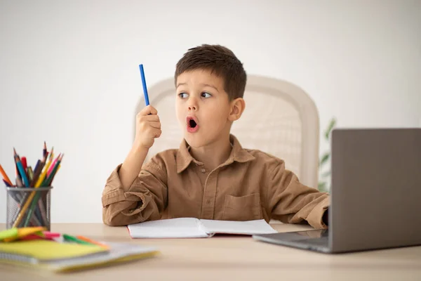 Happy surprised small european boy sit at table studying, drawing with laptop, looks at pencil with open mouth in room interior. Got idea, creation solution, art lesson and school, learning at home