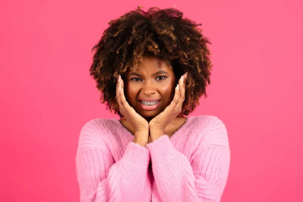 Sad dissatisfied smiling young black curly lady with braces in casual presses hands to cheeks, suffering from toothache, isolated on pink background, studio. Health care, dentistry, caries and pain