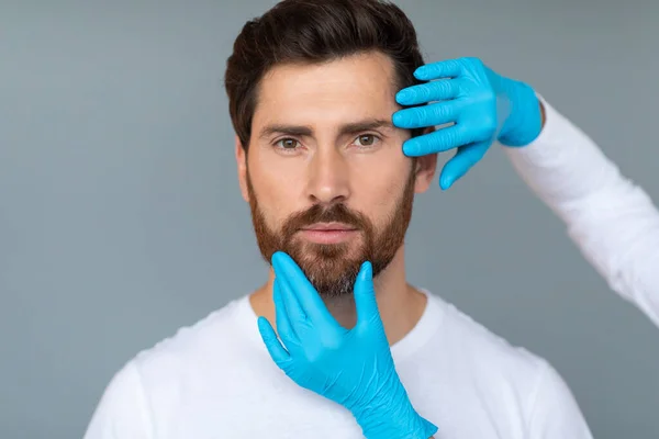 Plastic surgery and aesthetic cosmetology concept. Cosmetician hands in protective medical gloves touching male face over grey studio background