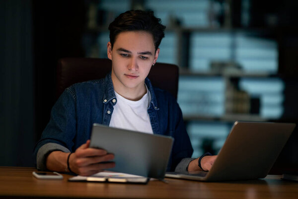 Concentrated young handsome man in casual outfit employee sitting at workdesk, using laptop and digital pad, working at office in evening, copy space. Modern technologies in business