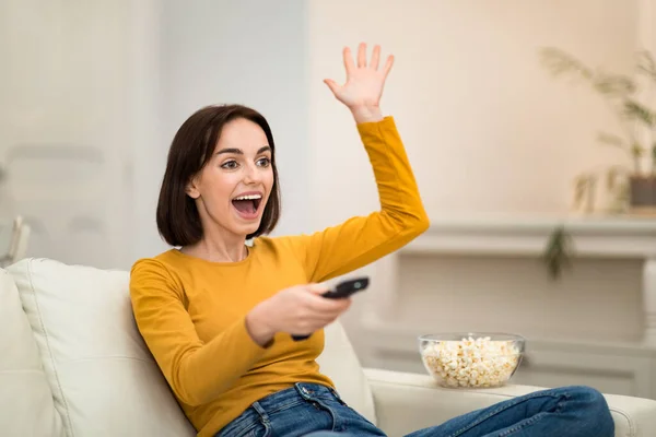 Emotional happy attractive young woman sitting on couch in cozy living room, watching TV show, football game with popcorn, holding remote controller, gesturing, spending time alone at home, copy space