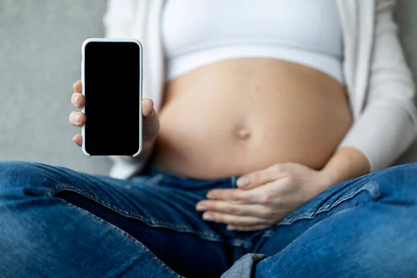 Unrecognizable Pregnant Woman Showing Blank Smartphone And Embracing Belly While Sitting On Couch At Home, Expectant Female Demonstrating Empty Cellphone With Black Screen, Cropped, Mockup