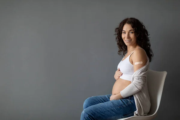 Portrait Of Smiling Young Pregnant Woman Posing After Coronavirus Vaccination, Expectant Female Sitting On Chair Over Grey Background, Touching Belly And Demonstrating Arm With Adhesive Bandage