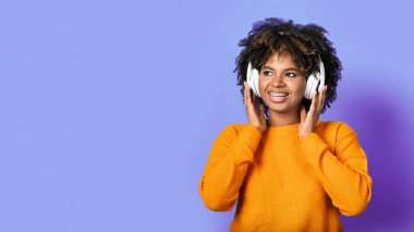 Closeup of positive cheerful pretty millennial black lady with bushy hair and teeth braces in orange sweater using wireless headphones, look at copy space for ad on purple studio background, panorama