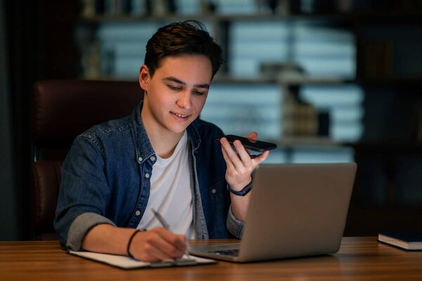 Hardworking young man manager staying late at dark office, motivated guy employee sitting at workdesk in front of laptop, recording voice mail on cell phone and taking notes, copy space